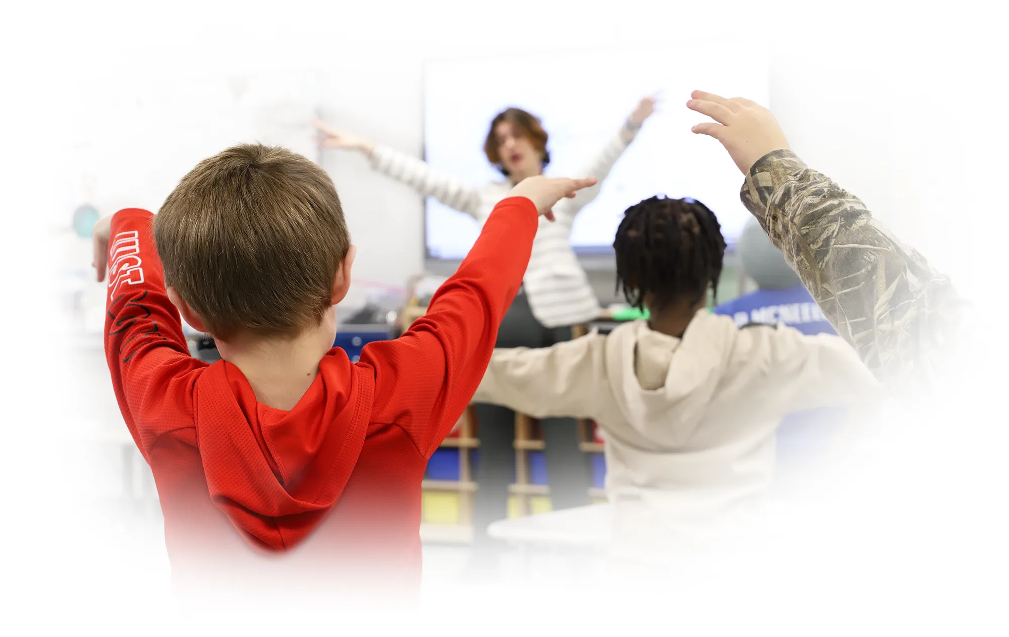 Children with their hands up in class while a teacher is instructing in the background