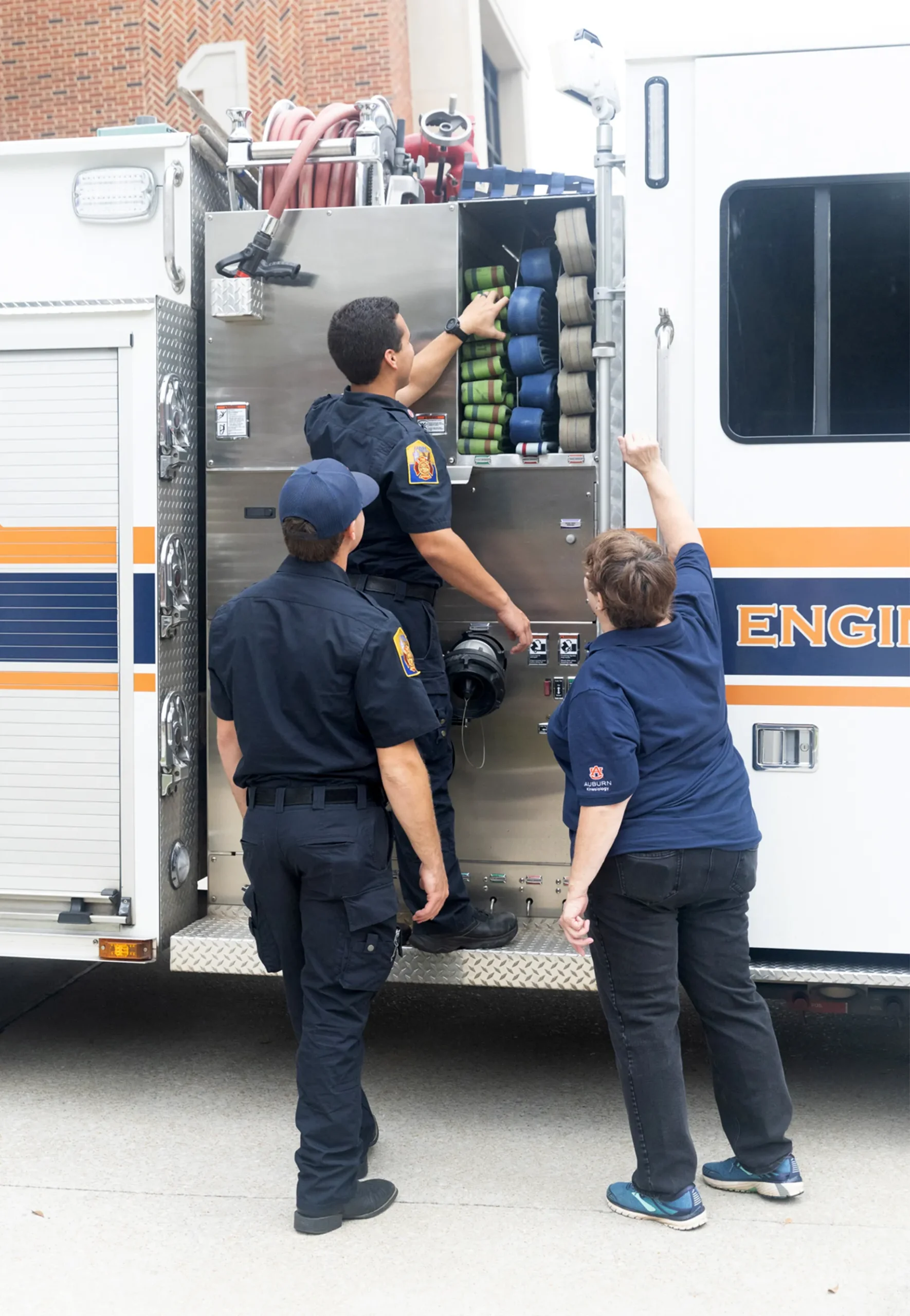 A firefighter in uniform shows equipment inside a fire truck to two colleagues, one of whom is pointing and another standing on the ground in a navy shirt with an Auburn University logo. Several neatly coiled hoses are visible inside the truck.