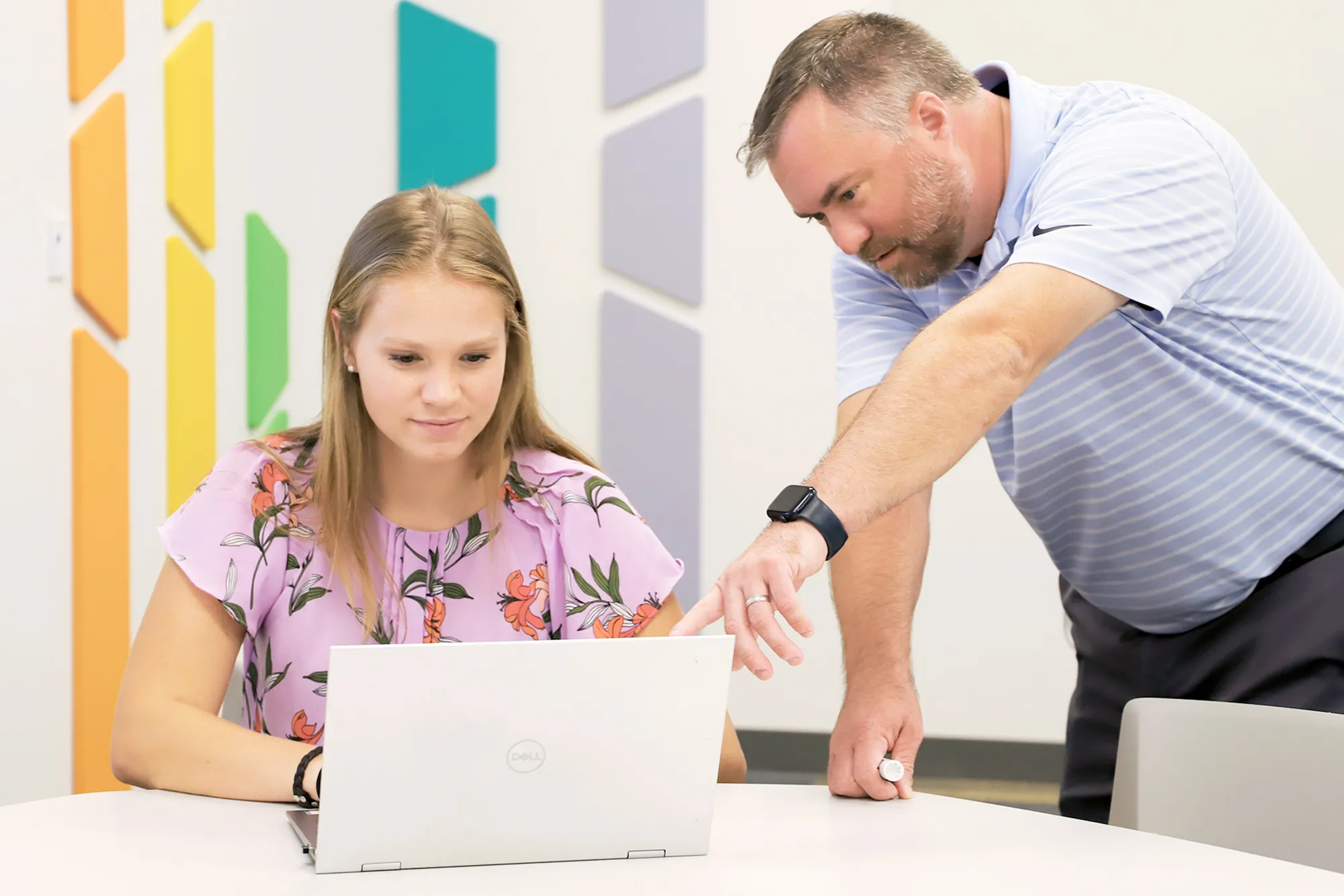 A young woman in a floral blouse works on a laptop while a man in a light blue striped shirt leans over and points at the screen, providing guidance. They are seated in a brightly decorated room with colorful geometric panels on the wall.