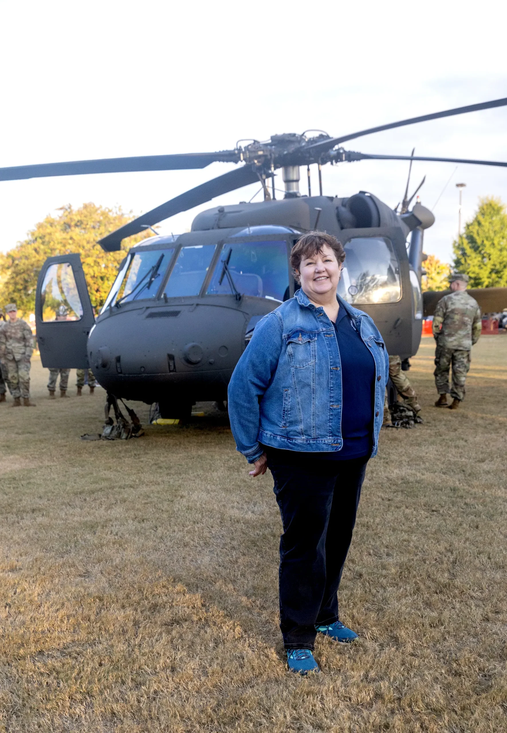 A woman wearing a denim jacket smiles while standing in front of a large military helicopter on a field. Army personnel are visible in the background near the aircraft.