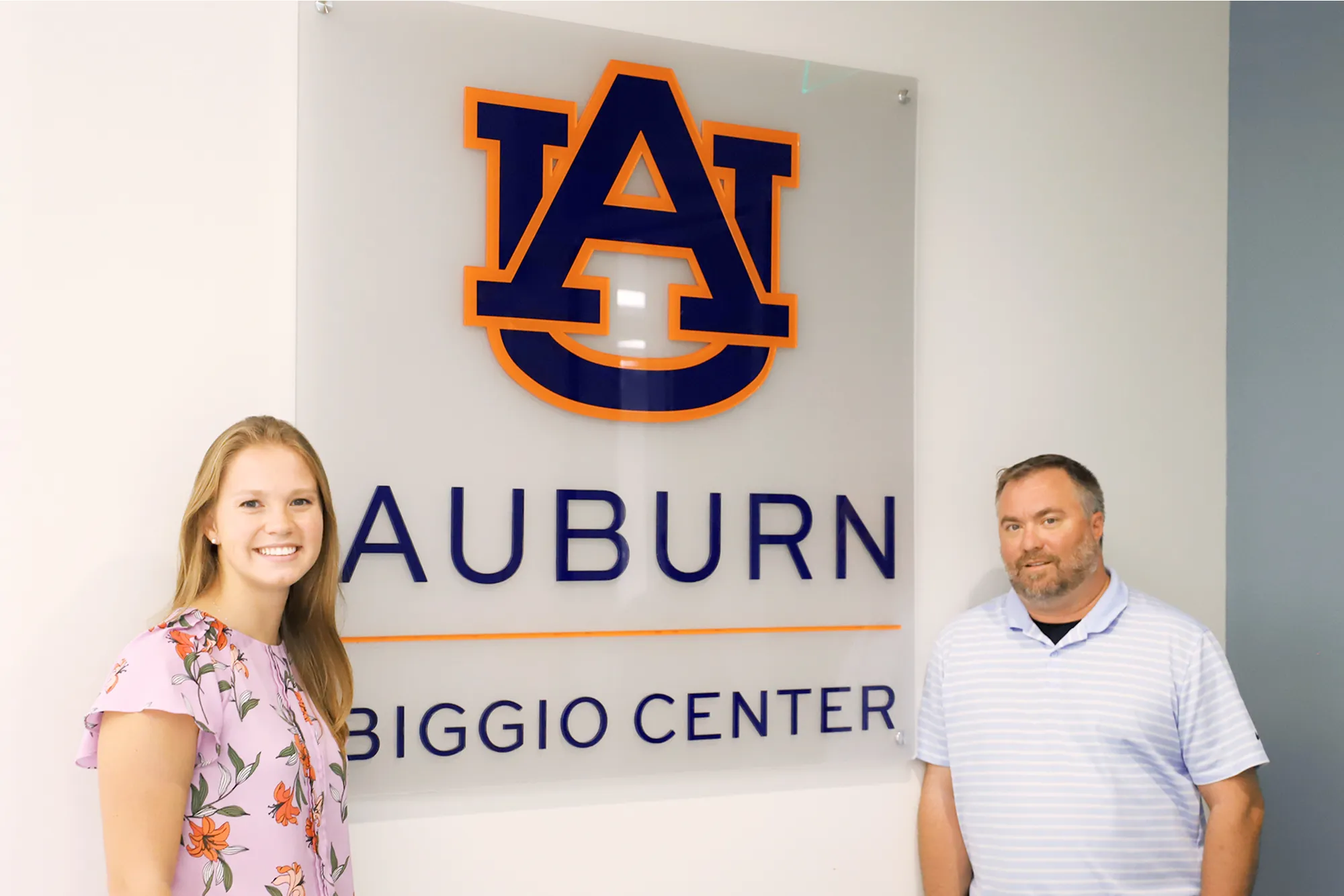A young woman and a man stand side by side, smiling in front of a wall sign that reads “Auburn Biggio Center” with the Auburn University logo displayed prominently above.