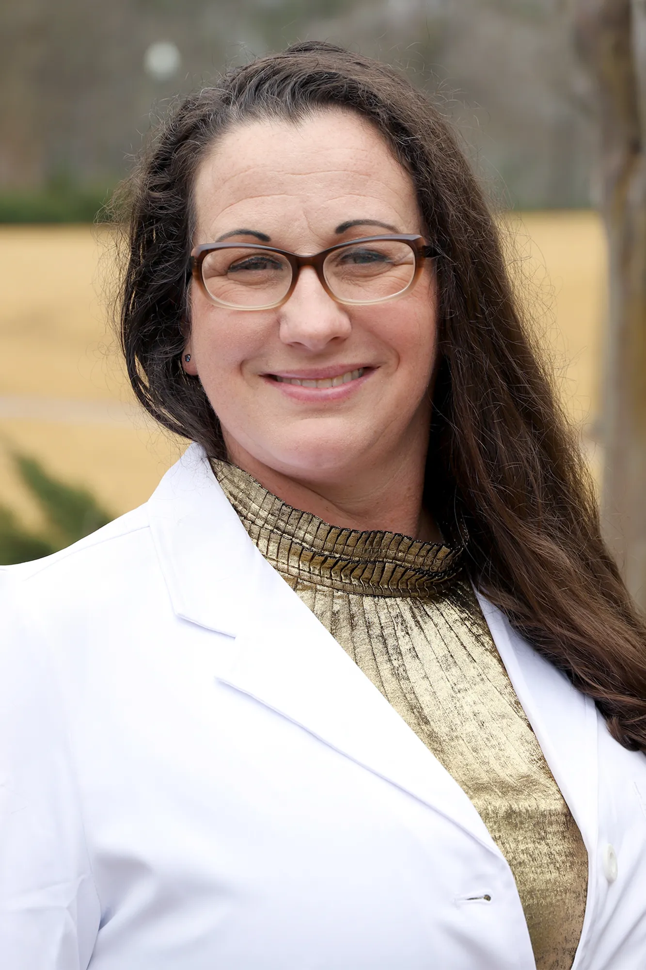 A woman with long dark brown hair and glasses smiles while wearing a white lab coat and a metallic gold top. The background is an outdoor scene with soft-focus trees and grass.
