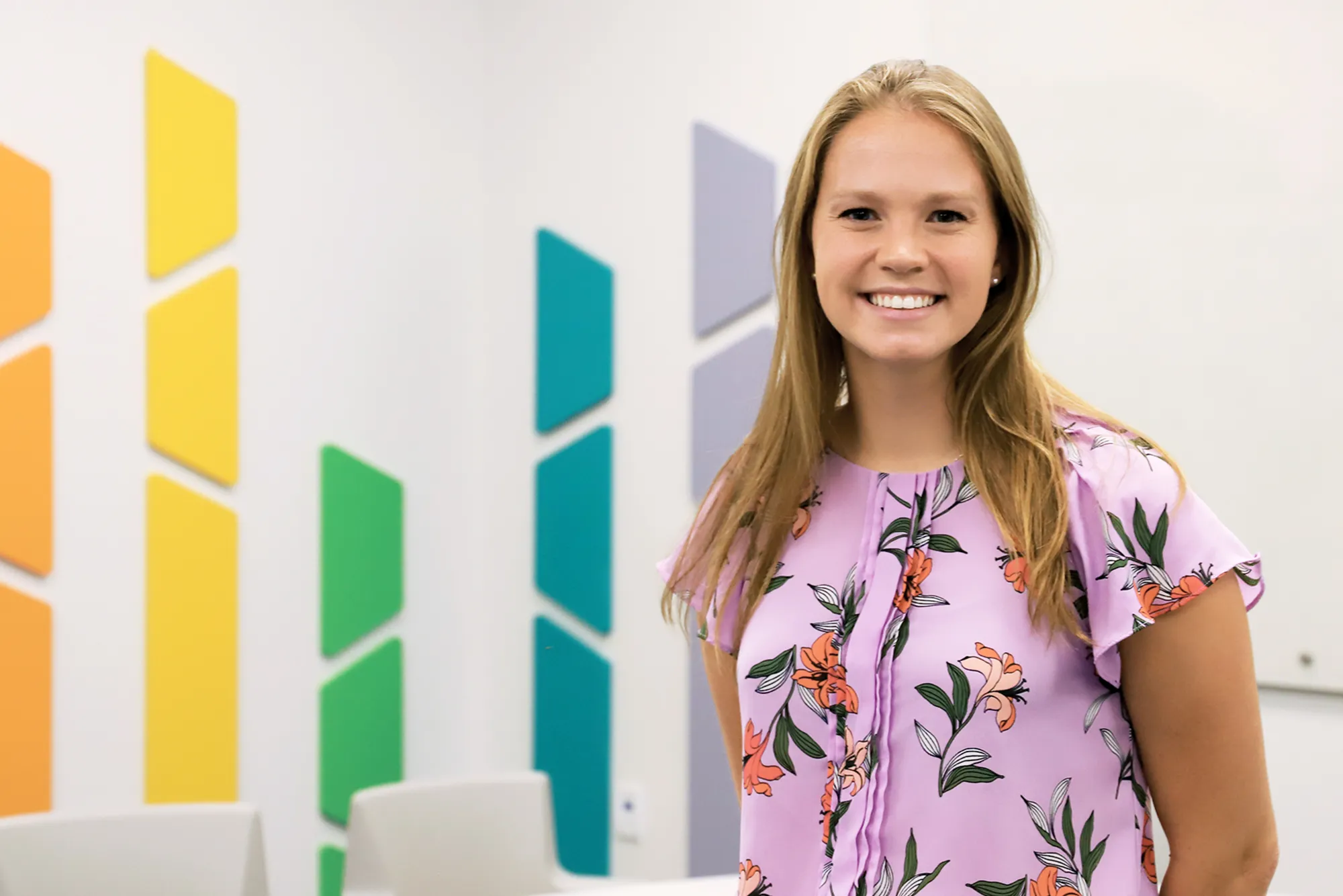 A young woman in a light purple floral blouse smiles at the camera while standing in a modern classroom with colorful geometric wall decorations in yellow, green, teal, and lavender.