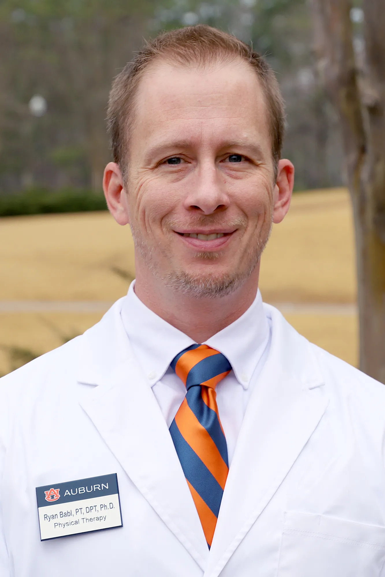 A man in a white lab coat with a name tag reading "Ryan Babl, PT, DPT, Ph.D., Physical Therapy" smiles outdoors. He wears an orange and blue striped tie and stands in front of a background with trees and grass.