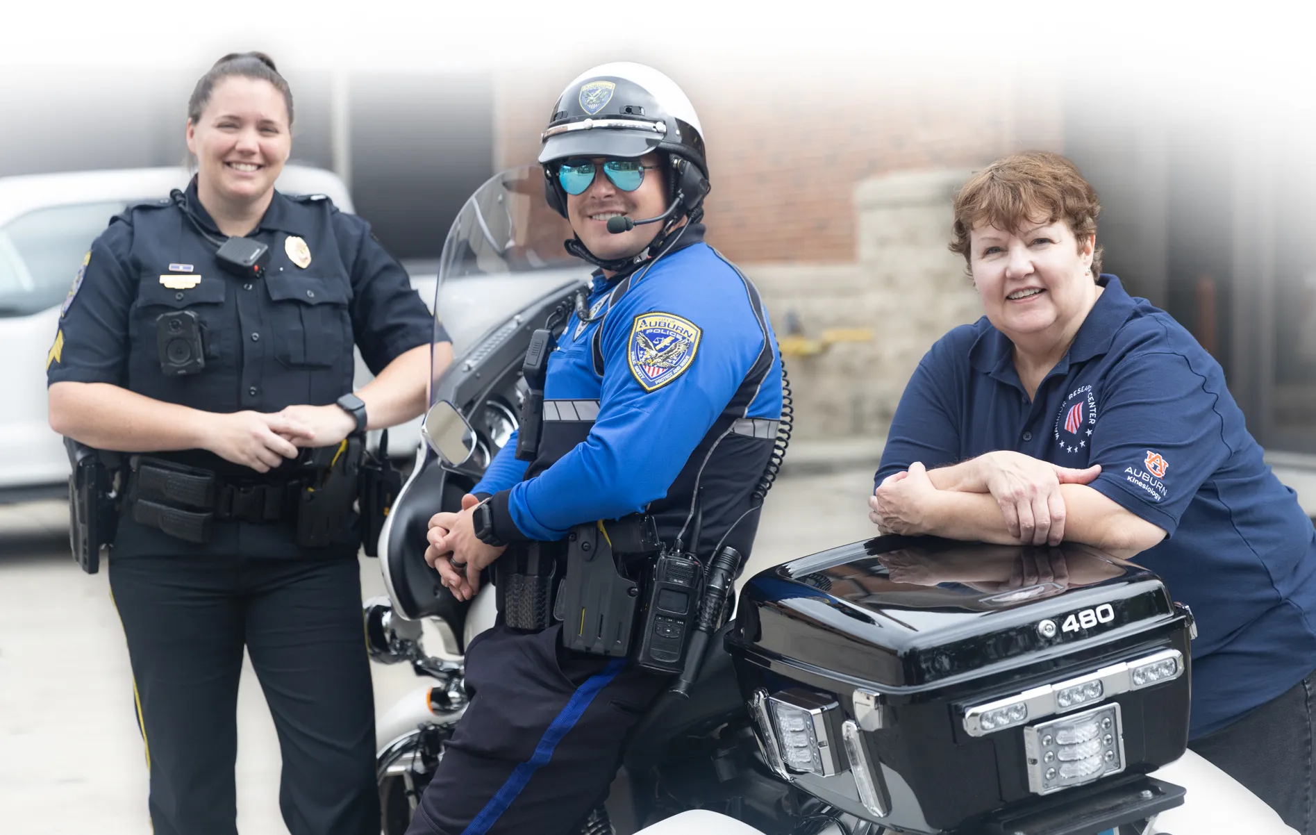 Three individuals pose with a police motorcycle—one officer in black uniform, one officer in a blue motorcycle patrol uniform with a helmet, and a woman in an Auburn University polo shirt. All are smiling outside a public safety building.