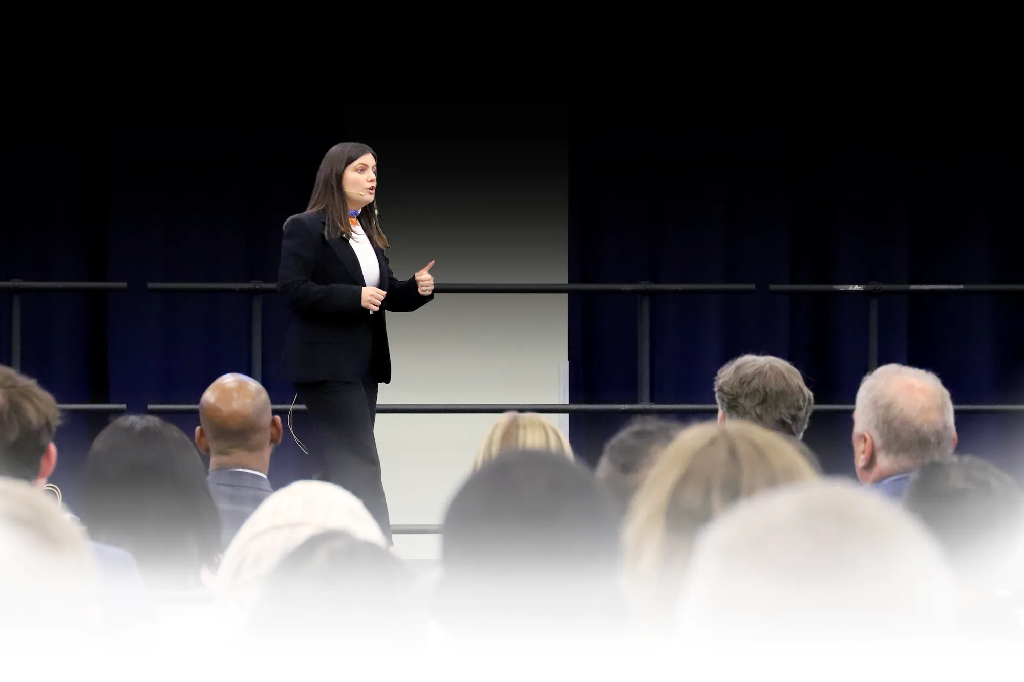 A woman in a black suit gives a presentation on stage in front of a seated audience. She is gesturing with her hand while speaking.