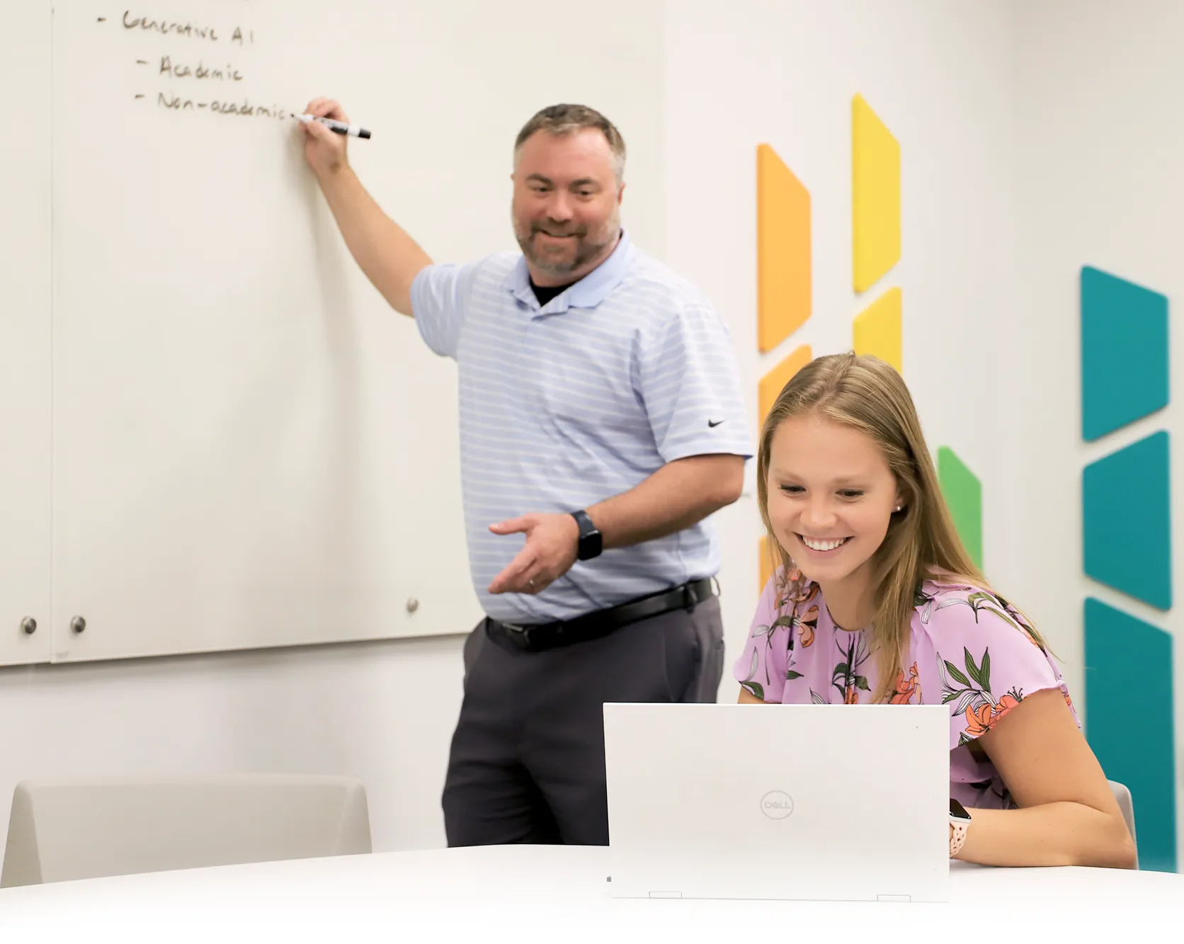 A man writes on a whiteboard labeled with topics like “Generative AI” while a young woman sits at a round table using a laptop. Both are smiling, and the classroom is decorated with colorful wall panels.
