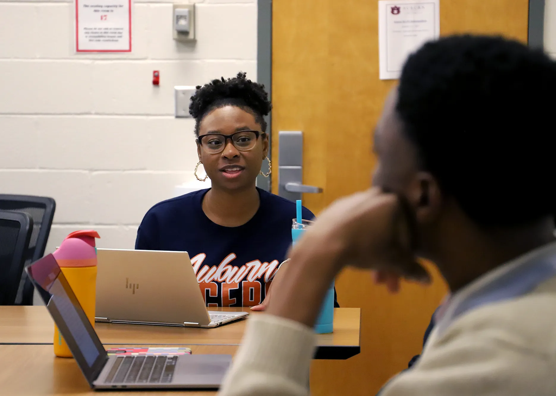 Female Auburn student wearing glasses and a navy “Auburn Tigers” shirt speaks during a classroom discussion, with an open laptop and water bottles on the table in front of her.