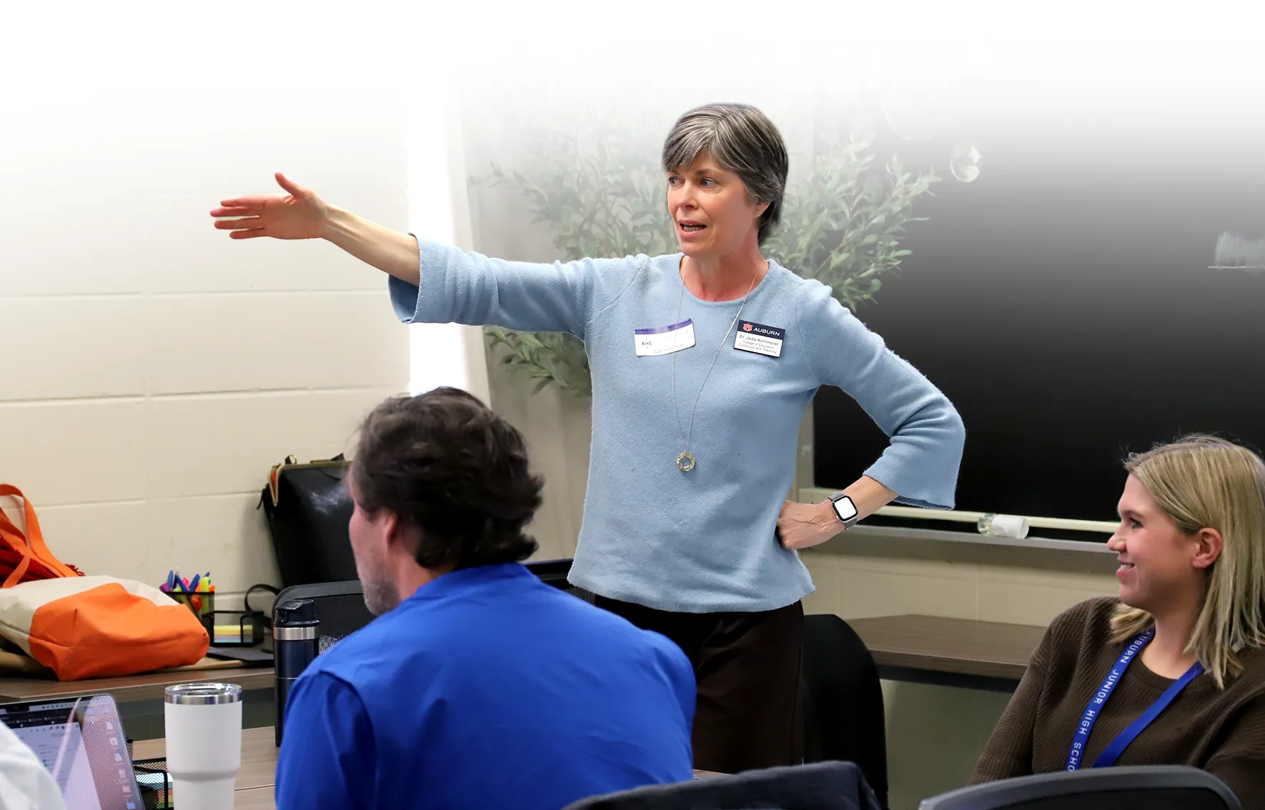 Instructor wearing a light blue sweater gestures with an outstretched arm while speaking to a group during an interactive classroom discussion at Auburn University.