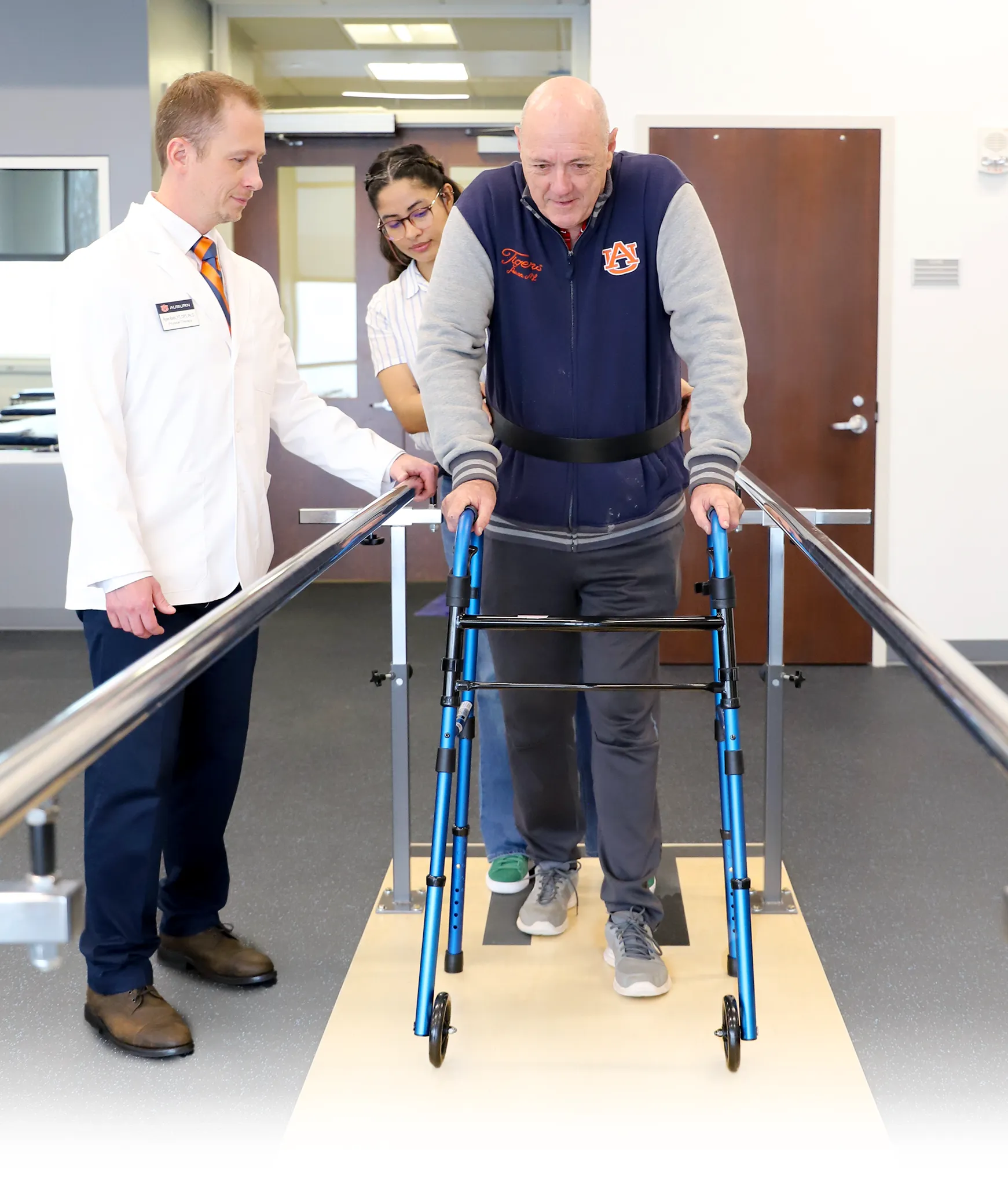A physical therapy patient uses a blue walker while walking between two parallel bars, assisted by a female therapist standing behind him. A male clinician in a white coat with an Auburn University name tag observes closely on the left. The patient wears an Auburn-branded jacket and is supported with a gait belt.