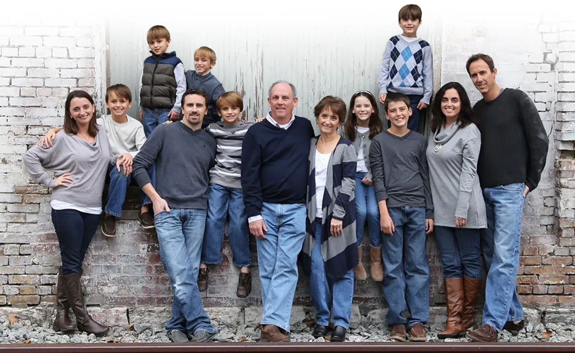 Large multigenerational family stands and sits together outdoors against a rustic white brick wall for a group portrait, including children and adults casually dressed in jeans and swea