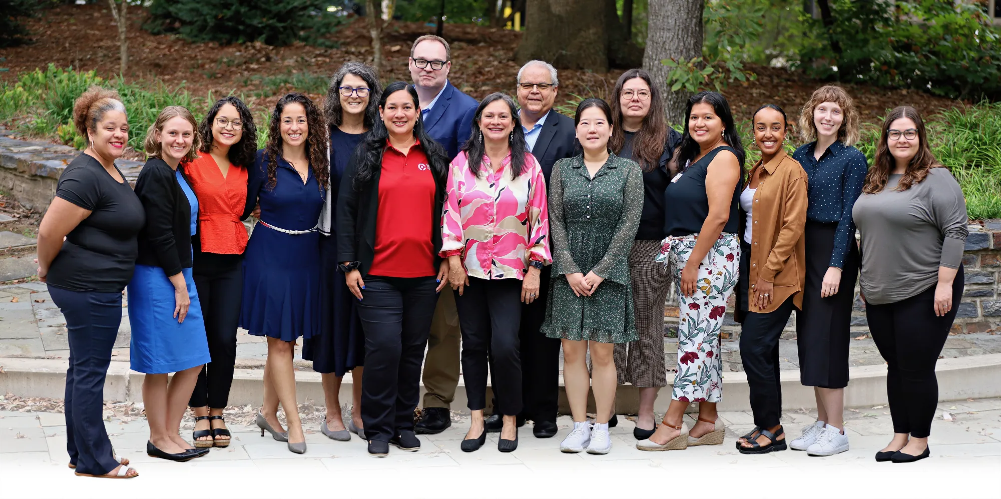 Diverse group of 17 adults, including men and women, pose together outdoors in business casual attire, standing on a stone path in front of trees and landscaped greenery.