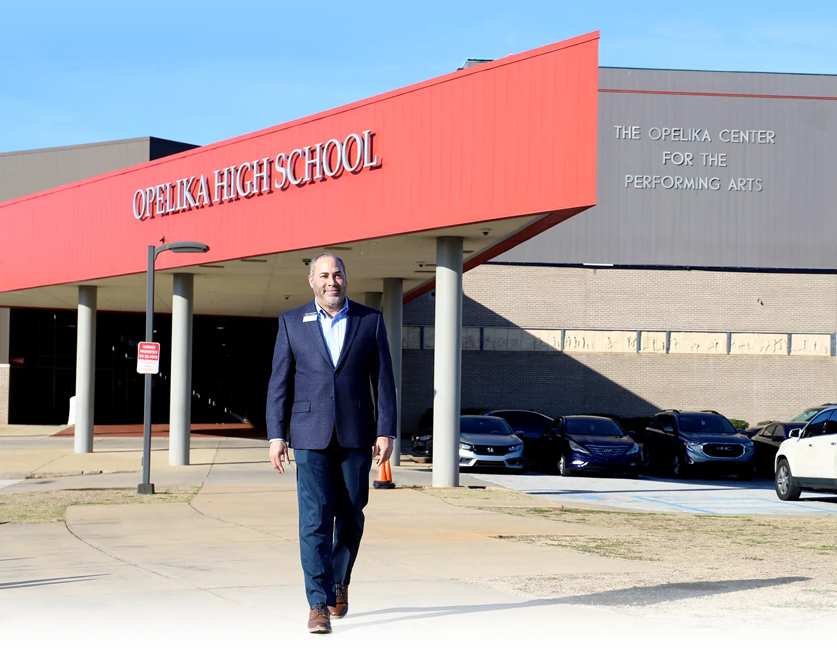 A man in a navy blazer walks confidently toward the camera outside Opelika High School. Behind him is a red overhang displaying the school name and the Opelika Center for the Performing Arts building. Several parked cars are visible in the background.