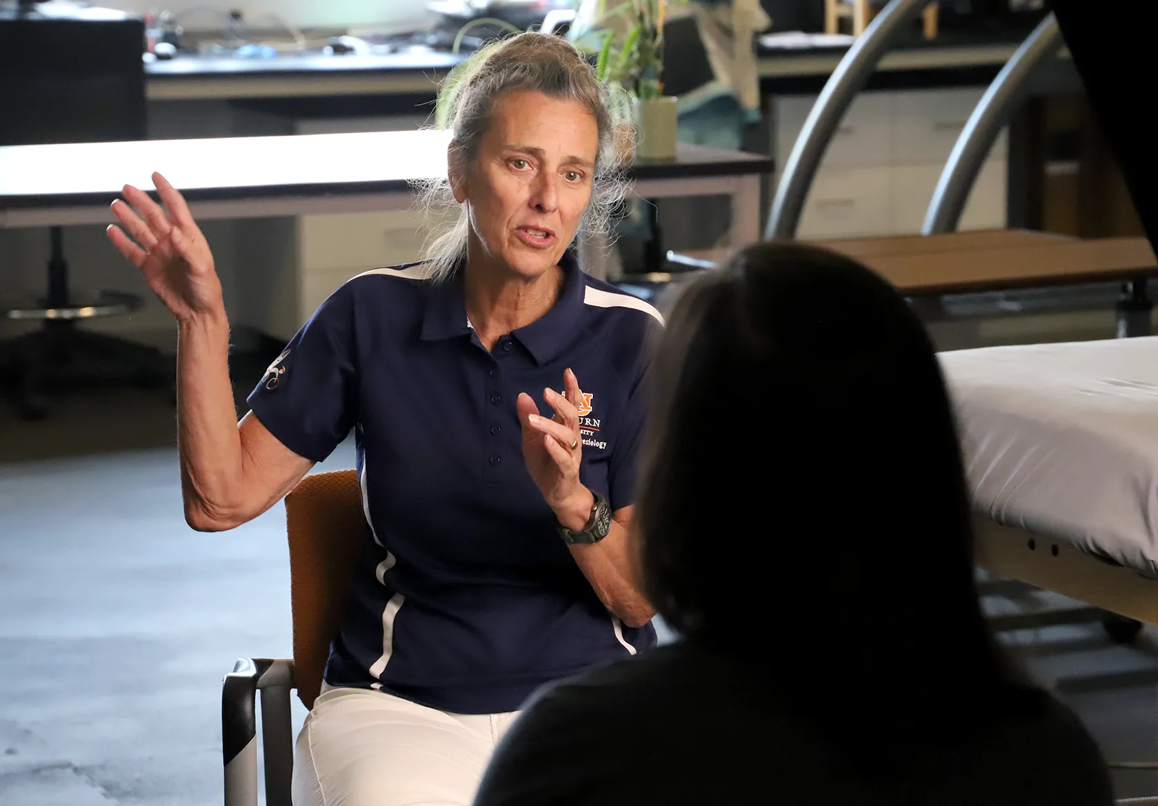 A woman in an Auburn University polo shirt sits in a laboratory-style room, gesturing with her hand while speaking to another person who is seen from behind. She appears engaged in conversation, with medical or research equipment visible in the background.