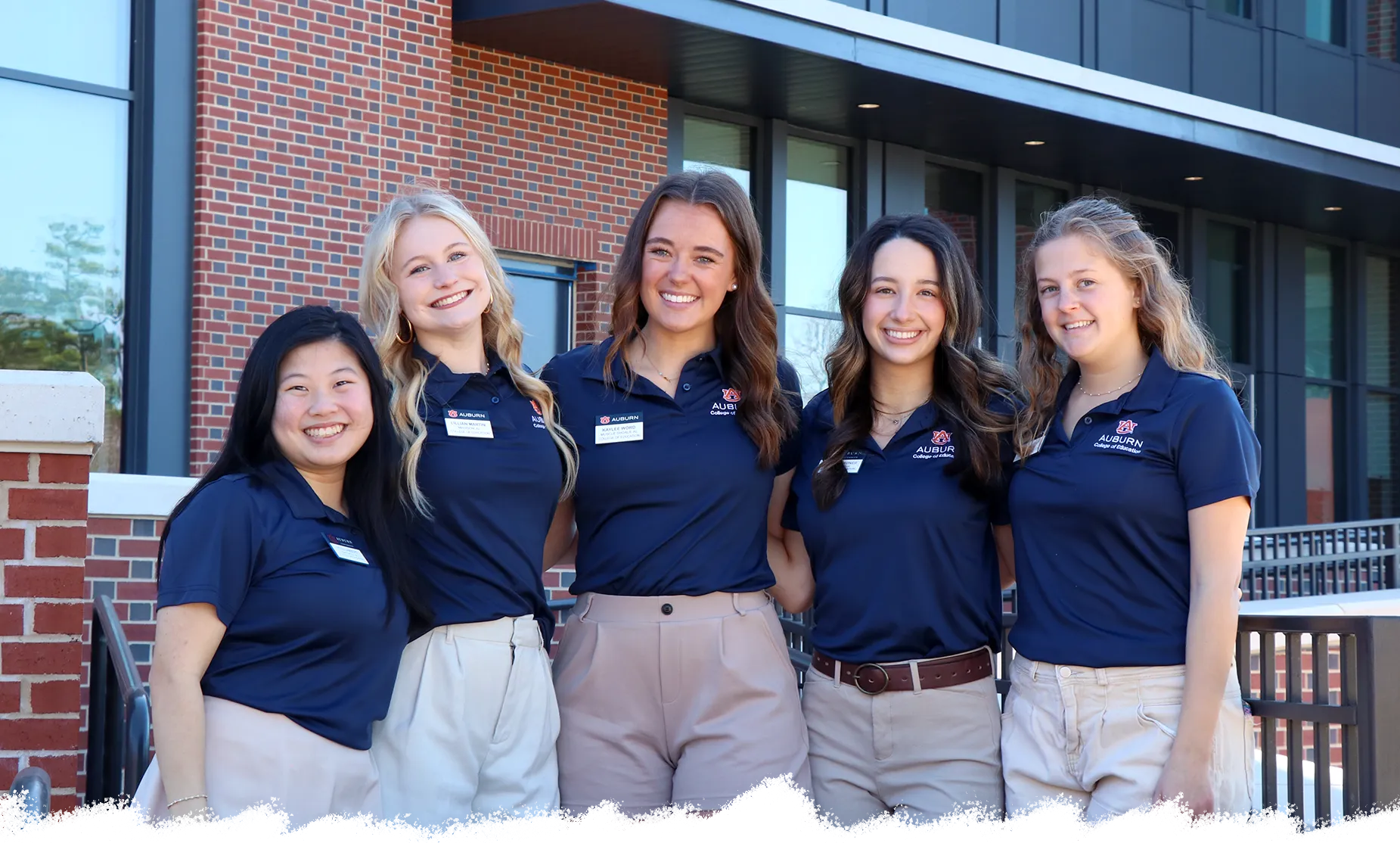 Five Auburn University students stand together outside a campus building, smiling and wearing matching navy blue College of Business polo shirts and khaki shorts or skirts. The background features modern brick and glass architecture, representing a welcoming and professional student leadership team.