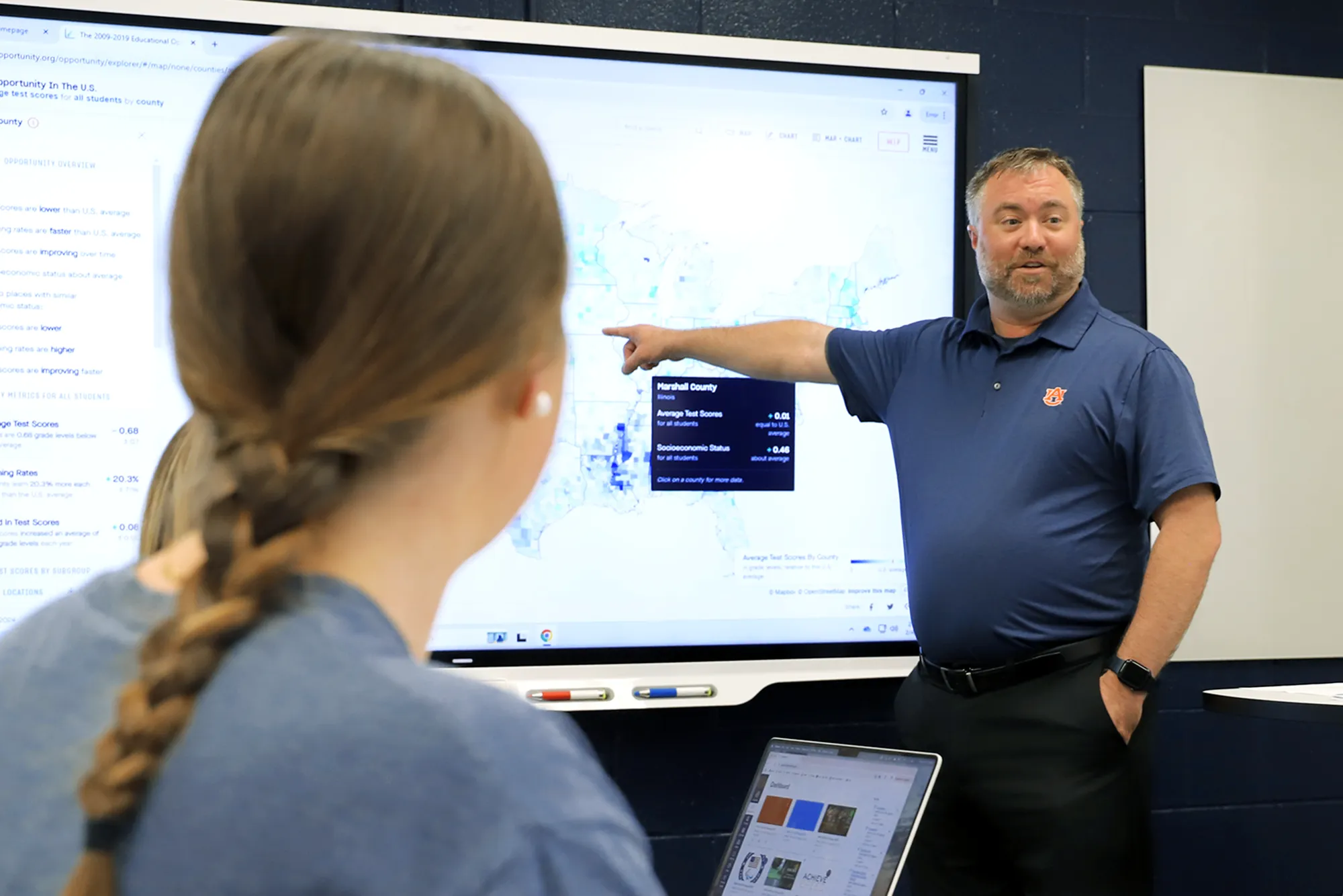 An Auburn University instructor in a navy polo shirt points at a large interactive map on a smartboard while teaching. A female student with a braid is seated in the foreground, watching attentively with a laptop open.