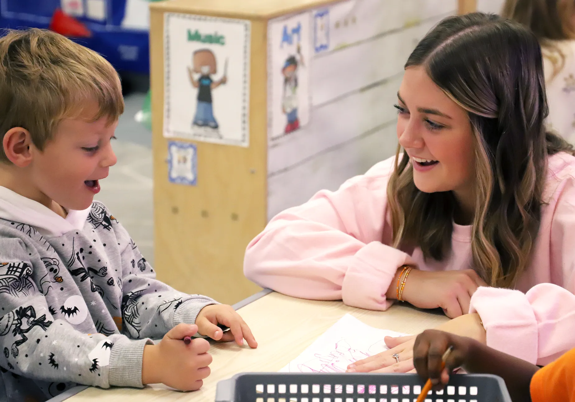 A young woman in a pink sweatshirt smiles while sitting at a table with a young boy who is holding a crayon and looking excited. They are engaged in a drawing activity in a brightly lit classroom with child-friendly artwork on the walls.