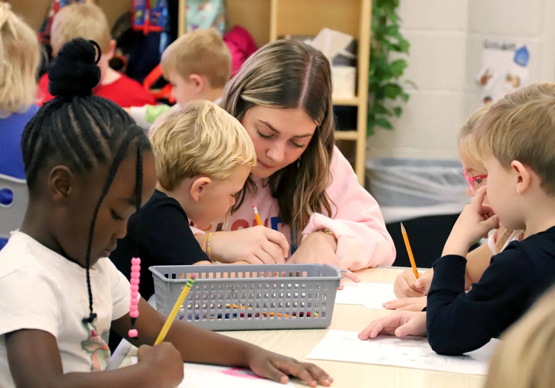 A young woman in a pink sweatshirt helps a young boy with his writing while other children work at the same table. One girl in the foreground concentrates on her drawing, and the classroom background is filled with students and colorful learning materials.
