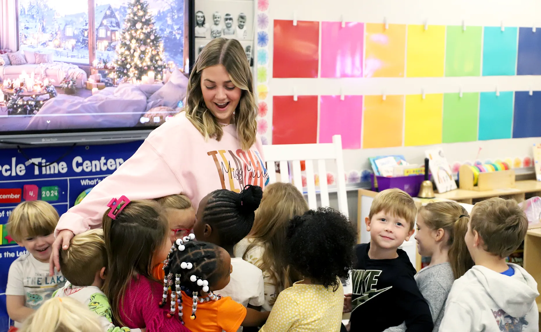 A teacher in a pink sweatshirt with the word “TEACHER” hugs a group of young children during a classroom activity. One child looks toward the camera smiling while others gather around the teacher. The colorful classroom features a rainbow-colored wall display and a digital screen showing a festive winter scene.