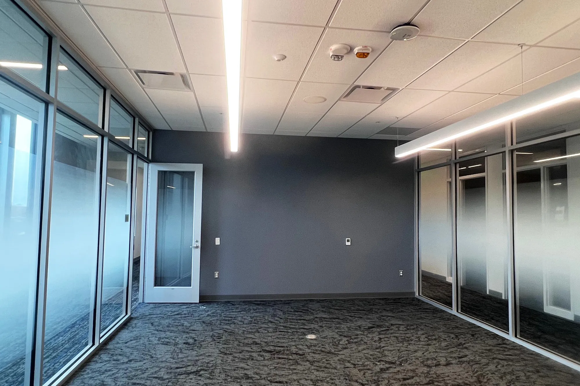Empty office or meeting space with a dark carpeted floor, glass walls on two sides, and modern linear ceiling lights inside the Auburn University College of Education building.