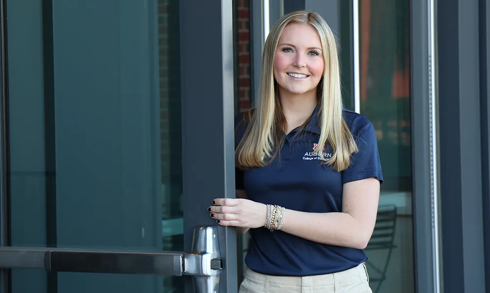 A smiling Auburn University student wearing a navy College of Education polo shirt and khaki shorts holds open a glass door, standing confidently in front of a modern building entrance.
