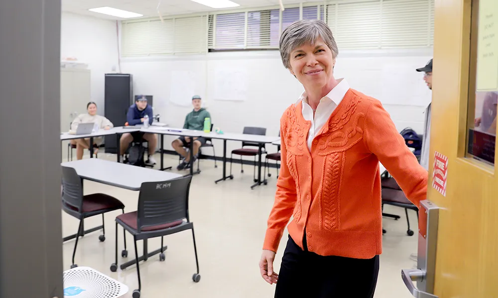 A female professor wearing an orange cardigan smiles as she enters a classroom, where several students are seated around a U-shaped arrangement of desks, suggesting a collaborative learning environment.
