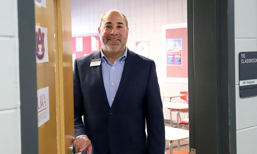 A male faculty member wearing a suit jacket and name tag stands smiling in the doorway of a classroom labeled 'Mr. Ferguson,' with Auburn-themed posters on the door and bulletin boards inside the room.