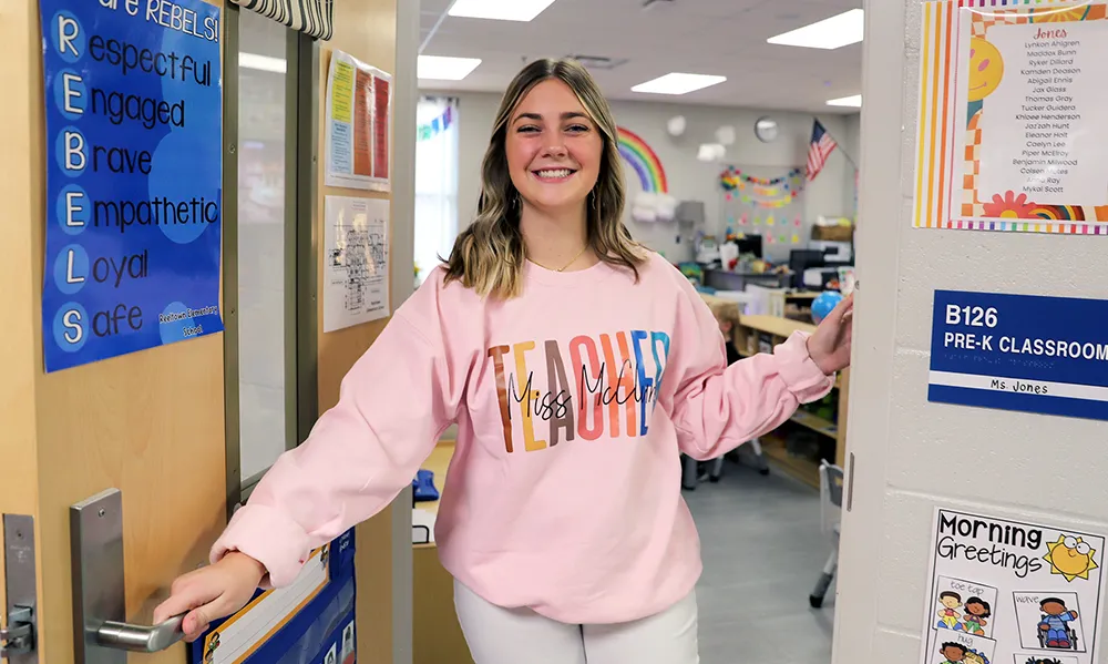 A young woman wearing a pink sweatshirt that says 'TEACHER Miss Molly' smiles while holding open the door to a colorful pre-K classroom labeled 'B126.' The room is decorated with rainbows and student artwork.