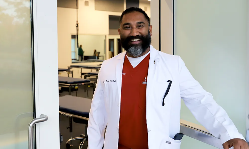 A man in a white lab coat and red scrubs stands smiling at the entrance to a clinical teaching space, with treatment tables and physical therapy equipment visible in the background.