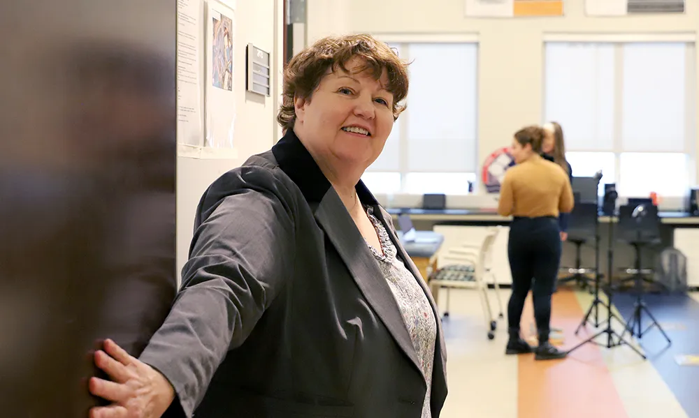 A woman with curly hair wearing a gray blazer stands near a classroom doorway, smiling at the camera. Inside the room, other individuals are gathered around musical instruments and camera equipment, indicating a performance or media activity.