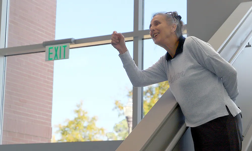 An older woman in a light gray sweater leans on a stair railing in a bright atrium with tall windows. She smiles and gestures expressively, with a green 'EXIT' sign and trees visible through the glass behind her.