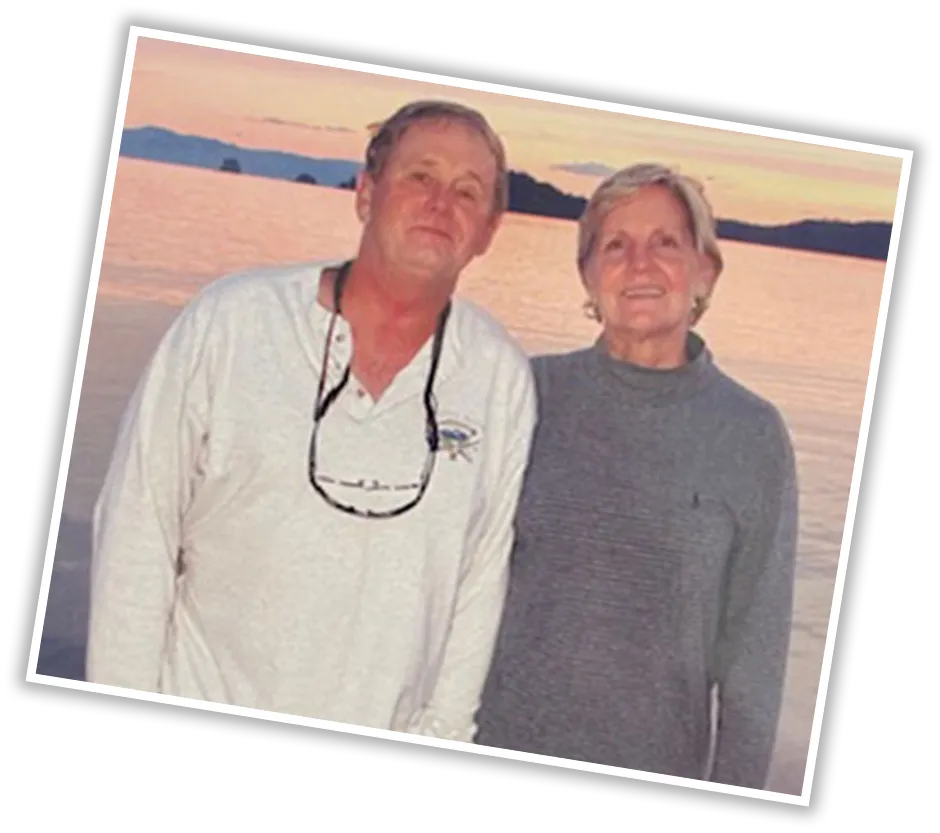 Candid outdoor photo of an older man and woman smiling together in front of a lake during sunset, with the man wearing a white long-sleeve shirt and the woman in a gray sweater.