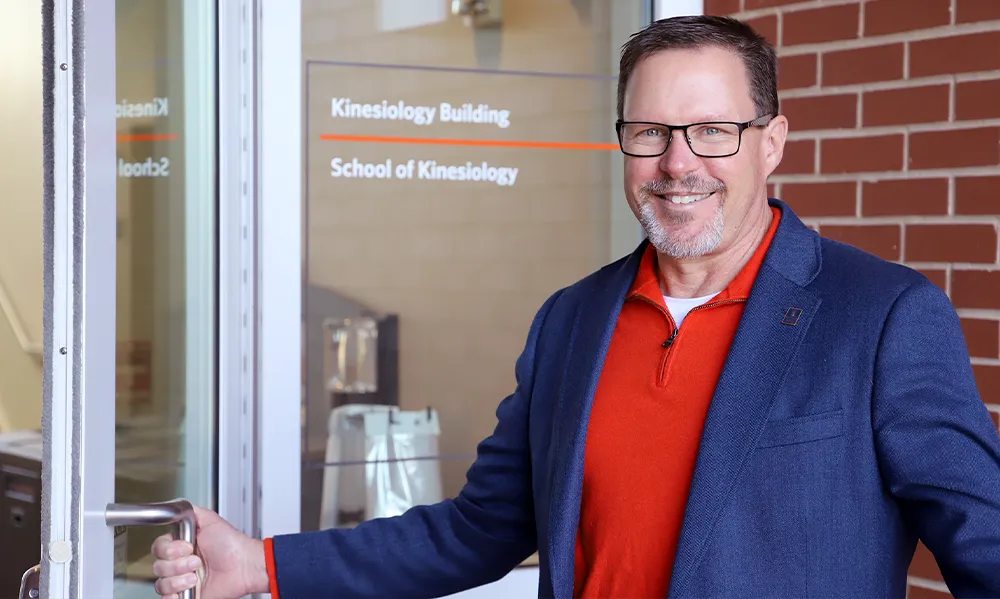 Dr. Jeffrey T. Fairbrother, Dean of Auburn University’s College of Education, stands smiling in front of the School of Kinesiology entrance. He wears glasses, a red zip-up shirt, and a navy blazer while holding open a glass door labeled “Kinesiology Building,” welcoming visitors to the facility.