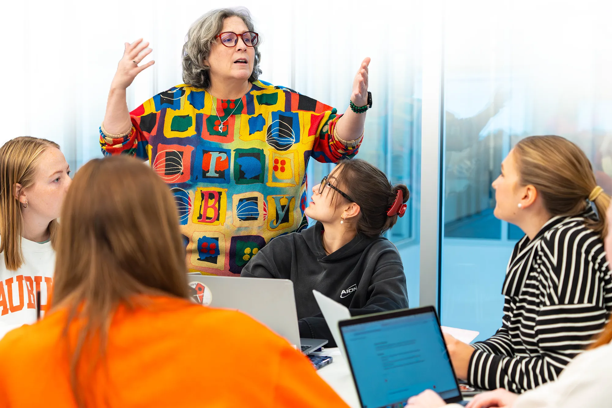Dr. Burton gestures while speaking to students gathered around a table with open laptops during class discussion.