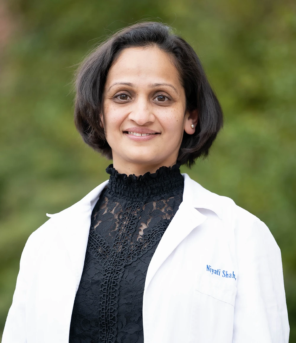 Portrait of Niyati Shah in a white coat and black top against a softly blurred green outdoor background.