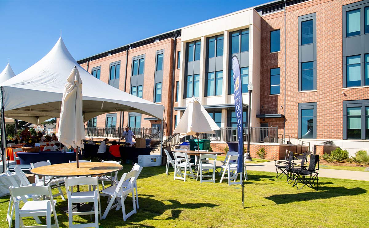 Exterior view of the brick College of Education building with white event tents, round tables, and white chairs set up on a green lawn under a clear blue sky.