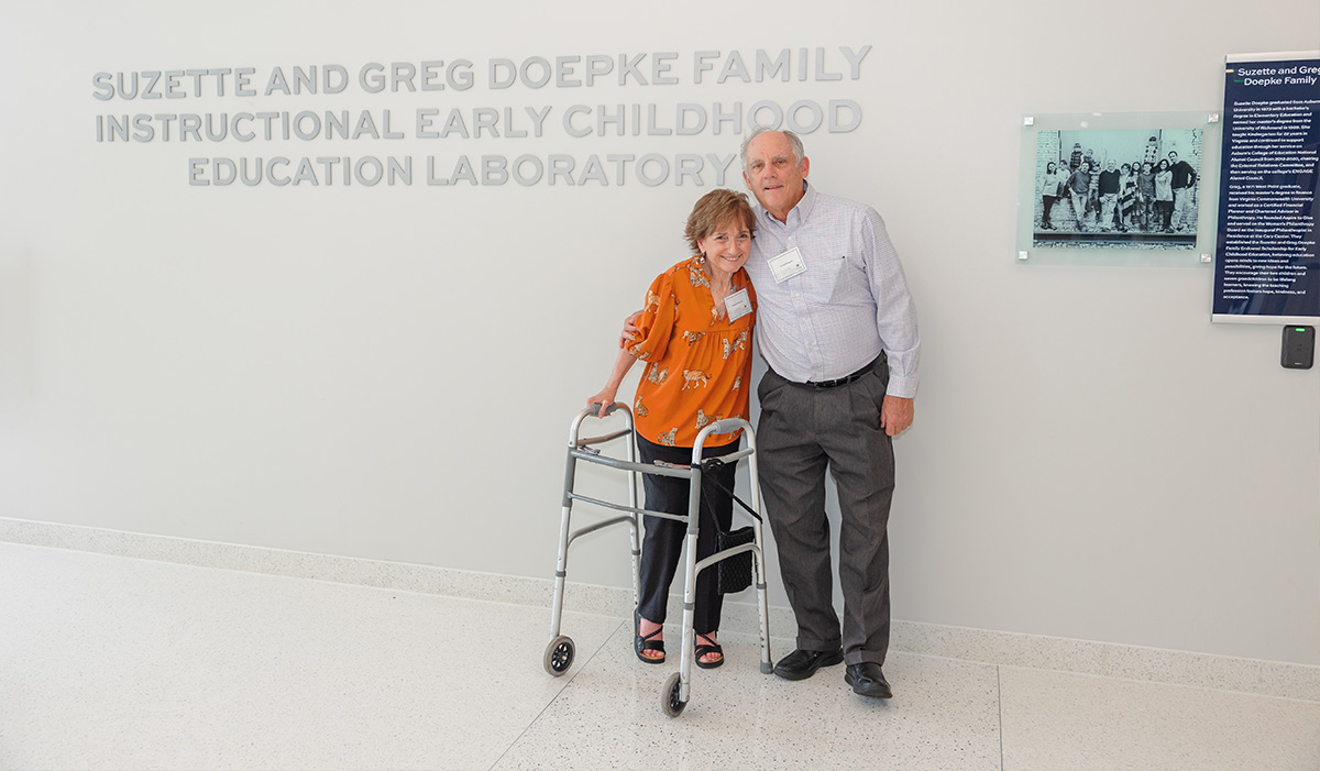 Suzette and Greg Doepke stand together in a hallway next to a wall with silver lettering: Suzette and Greg Doepke Family Instructional Early Childhood Education Laboratory.