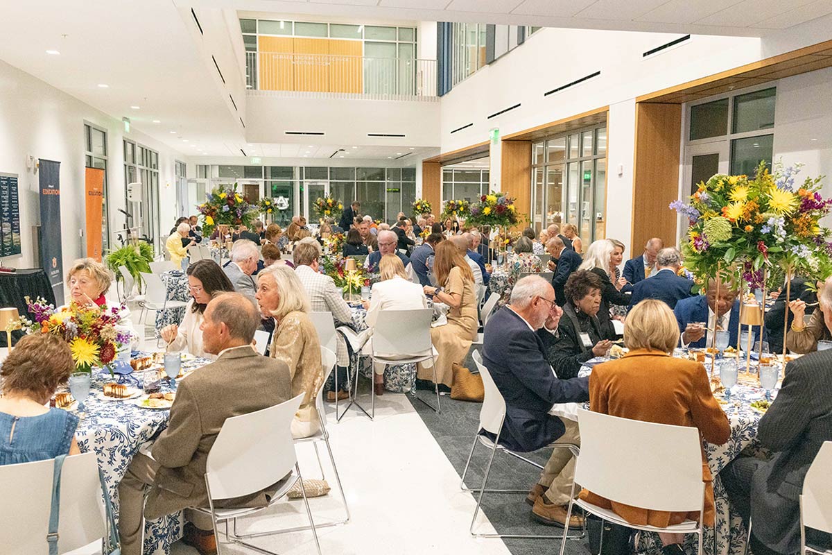 High-angle view of a crowded formal dinner in a large indoor atrium. Guests sit at round tables with blue patterned cloths and floral centerpieces.