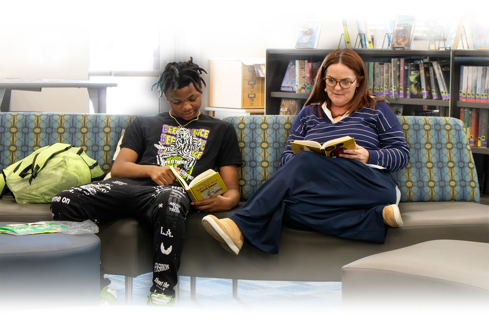 Student and instructor read books together on a couch in a school library during a literacy activity.