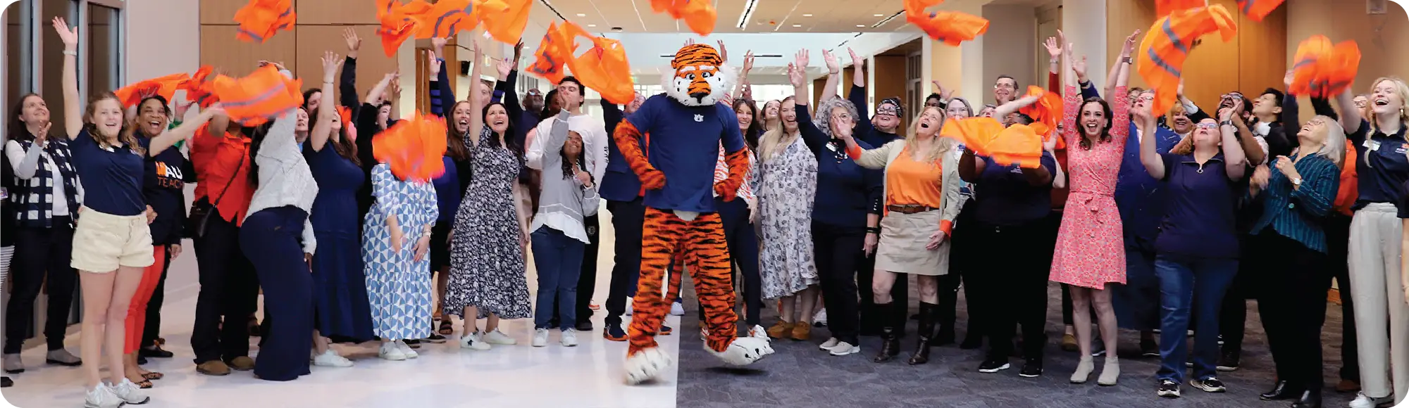 Aubie stands with Auburn College of Education faculty and staff as they celebrate indoors, tossing orange rally towels into the air.