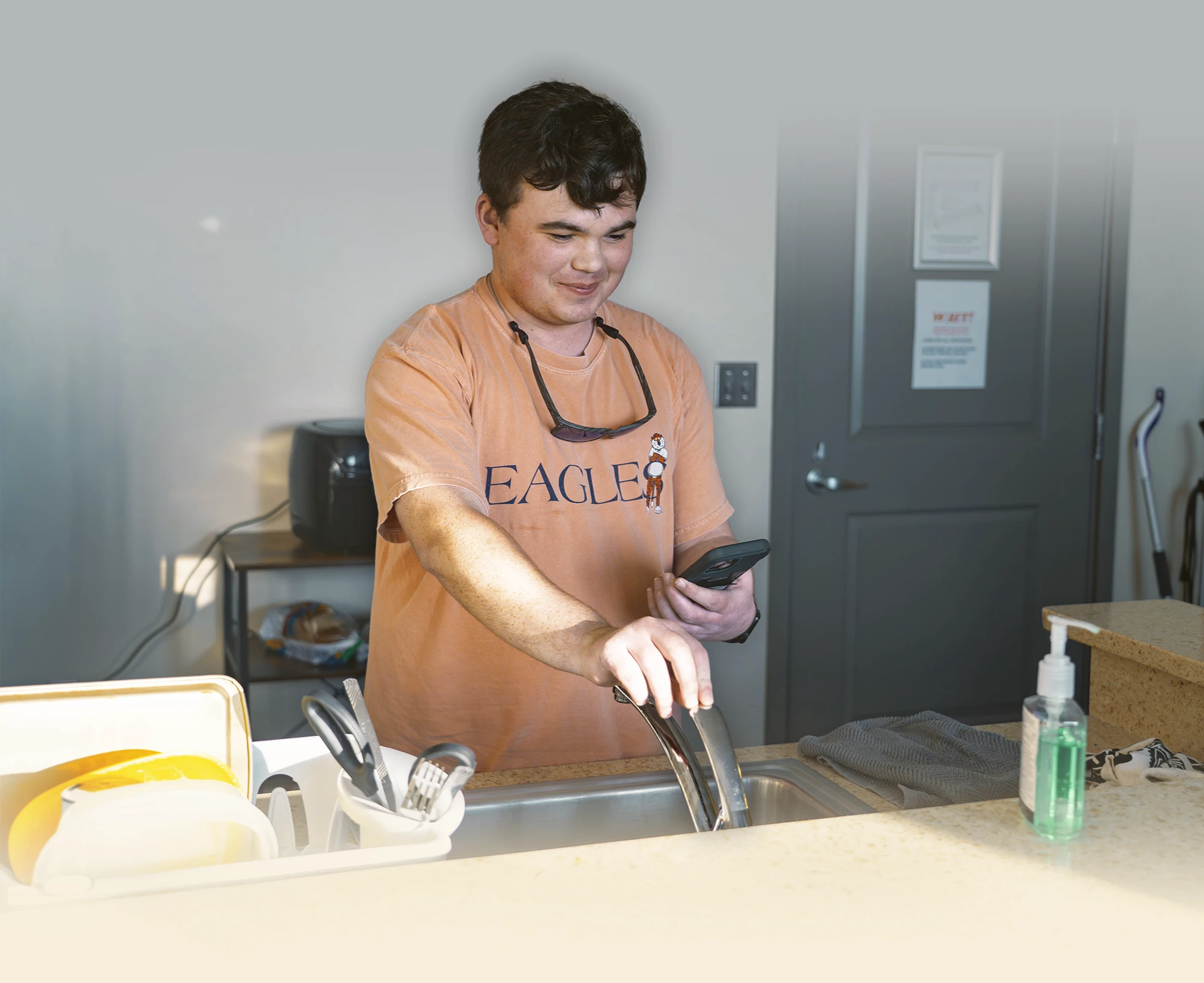 Student stands at a kitchen sink holding a phone while turning on the faucet in an apartment.